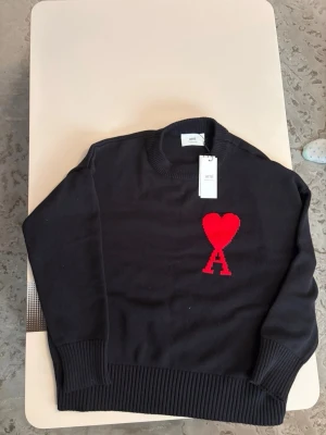 Svart stickad tröja från Ami Paris - Svart stickad tröja från Ami Paris med röd hjärtlogga och bokstaven A på bröstet. Tröjan har rund halsringning, ribbade muddar och nederkant. Perfekt för dig som gillar stilrena plagg med en twist.