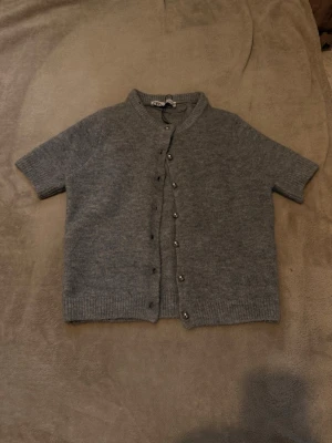 Grå kortärmad kofta från ZARA - Säljer en grå kofta från ZARA med korta ärmar och rund hals. Koftan har ribbstickade muddar och knappar framtill i silverfärg. Materialet ser ut att vara mjukt och stickat, perfekt för lager-på-lager och en chill look.