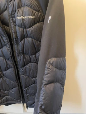 Svart pufferjacka Peak Performance - Snygg svart pufferjacka från Peak Performance med quiltat mönster och dragkedja framtill. Jackan har huva, mjuka ärmar i stretchmaterial och Peak Performance-logga broderad på bröstet. Perfekt för vår/höst och riktigt skön att bära. Storlek S. Bra skick!