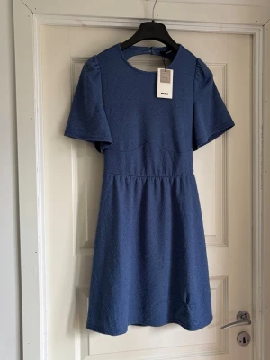 Blå klänning från Bik Bok - Supersöt blå klänning från Bik Bok i återvunnen polyester. Klänningen har korta, vida ärmar, rund halsringning och markerad midja. Materialet har en strukturerad yta som ger en cool känsla. Perfekt för dig som gillar stilrena och trendiga plagg.