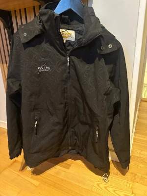 Svart vindjacka från On The Peak storlek 38 - Fin och hel. Enda är att gummibandet nertill på jackan har börjat ”torka” så man kan inte justera den så enkelt nertill. Inget som påverkar själva jackan dock. Snygg svart vindjacka från On The Peak med diskret blommönster, huva och dragkedja framtill. Två fickor med dragkedja och justerbar nederkant. Perfekt för blåsiga dagar och enkel att matcha med olika outfits.