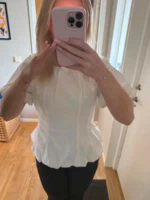 Blus  - Söt vit topp i krispigt material med fin struktur och lätt puffiga kortärmar. Figursydd passform med markerad midja och diskret volang nertill som gör den extra piffig.Perfekt till både jeans för en avslappnad look eller kjol för något mer uppklätt 🤍 Dragkedja bak. Aldrig använd så i mycket fint skick!  📦 Skickas snabbt 💬 Skriv vid frågor eller fler bilder