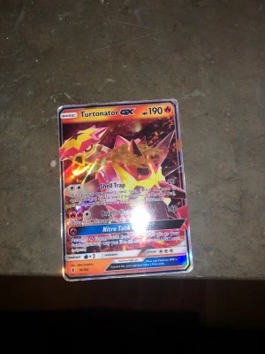 Turtonator GX Pokémonkort - Säljer ett Turtonator GX samlarkort från Pokémon med holografisk yta. Kortet har färger i rött, gult och orange med coola effekter och attacker som Shell Trap, Bright Flame och Nitro Tank GX. Perfekt för dig som gillar att samla eller spela Pokémonkort.