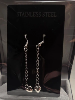 Silverfärgade örhängen med hjärtan - Säljer ett par stilrena silverfärgade örhängen i rostfritt stål. De har en tunn kedja och längst ner hänger små blanka hjärtan. Perfekta för dig som gillar minimalistisk och söt design. Krokarna är enkla att använda och har plaststopp på baksidan. Längden är ca 4 cm.