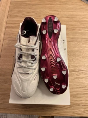 Puma King Platinum fotbollsskor - Snygga Puma King Platinum fotbollsskor i vitt och vinrött med metallicröd sula och klassisk design. Ovandelen är i mjukt läder och mesh för bra komfort och känsla på planen. Skorna har fasta dobbar och ikoniska King-detaljer. Perfekta för dig som vill sticka ut på planen.