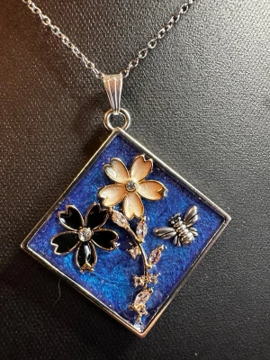 Halsband med blått fyrkantigt hänge - Unikt halsband med ett fyrkantigt hänge i blått, dekorerat med två blommor i svart och vitt samt en liten silverfärgad bi. Hänget har gnistrande stenar i mitten av blommorna och på stjälken. Kedjan är silverfärgad och smal, perfekt för att ge en cool touch till din outfit.