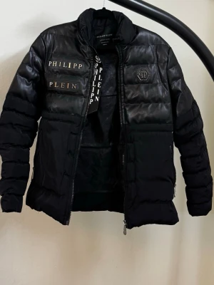 Svart pufferjacka från Philipp Plein - Svart pufferjacka från Philipp Plein med glänsande partier upptill och tydliga logodetaljer på bröstet. Jackan har hög krage, dragkedja framtill och diskreta dödskalletryck på innerfodret. Perfekt för kalla vinterdagar och riktigt snygg streetstil.
