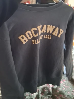 Svart sweatshirt Rockaway Beach - Mörk sweatshirt med trycket 'ROCKAWAY BEACH NY 1999' i beige på bröstet. Tröjan har rund halsringning och långa ärmar. Tillverkad i mjukt material, perfekt för en avslappnad stil. Klassisk och enkel design utan huva.