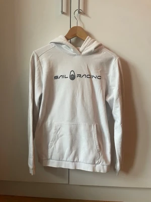 Vit hoodie från Sail Racing - PRIS KAN DISKUTERAS. Vit hoodie från Sail Racing med svart logga och tryck på bröstet. Klassisk känguruficka framtill och huva med tryckt detalj. Tillverkad i mjukt bomullsmaterial som känns skönt mot huden. Perfekt för en avslappnad och sportig stil.