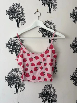 Rosa croppad linnetopp med jordgubbar - Söt rosa croppad linnetopp med tunna axelband och ett lekfullt jordgubbsmönster. Perfekt för dig som gillar färgglada och roliga plagg. Toppen har en lätt rynkad nederkant och ärmlös design som passar till sommarens outfits.