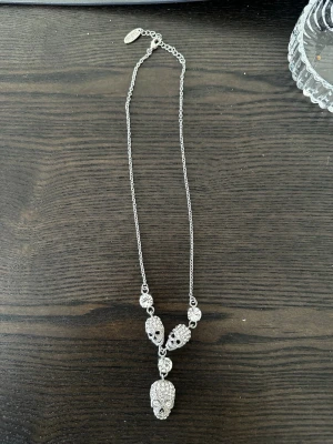 Rock by Sweden halsband  - Unikt halsband i silverfärg med flera döskallehängen dekorerade med gnistrande stenar. Kedjan är tunn och har ett litet märke vid låset. Perfekt för dig som gillar edgy och coola accessoarer.