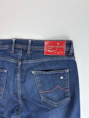 Mörkblå jeans från Jacob Cohen - Snygga mörkblå jeans från Jacob Cohen med raka ben och klassisk femficksdesign. Jeansen har röda kontrastsömmar, en röd patch med logga bak och en liten blomdetalj på bakfickan. Tillverkade i mjuk denim med stretch för extra komfort.
