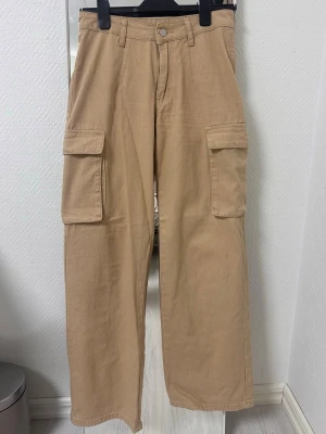 Beige cargobyxor med stora fickor - Stilrena beige cargobyxor med raka ben och två stora fickor på sidorna. Byxorna har hög midja och stängs med knapp och dragkedja. Tillverkade i ett mjukt bomullstyg som ger en avslappnad och trendig look.