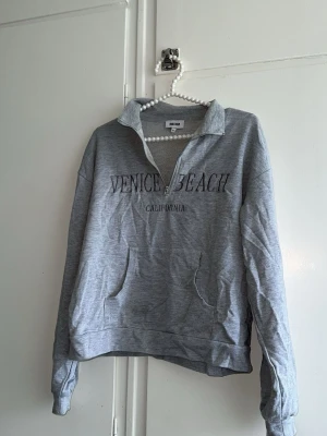 Grå quarterzip Venice Beach-tröja - Snygg grå sweatshirt med quarterzip och broderad text 'Venice Beach California' på bröstet. Tröjan har en hög krage, känguruficka framtill och är gjord i mjukt bomullsmaterial. Perfekt för en avslappnad och trendig look.