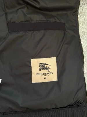 Svart jacka från Burberry, M - Snygg svart jacka från Burberry med klassiskt rutigt foder i beige och svart. Jackan har dragkedja och ribbade muddar. Perfekt för dig som vill ha en stilren och tidlös look med exklusiv känsla.