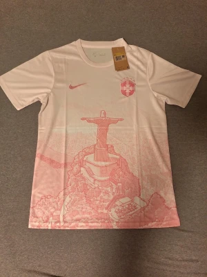 Rosa Brasilien Nike fotbollströja - Vit och fotbollströja från Nike med Brasilien-emblem och rosa detaljer. Framsidan har ett stort tryck av Kristusstatyn i Rio de Janeiro i rosa. Tröjan har rund hals och korta ärmar, tillverkad i lätt och ventilerande material.