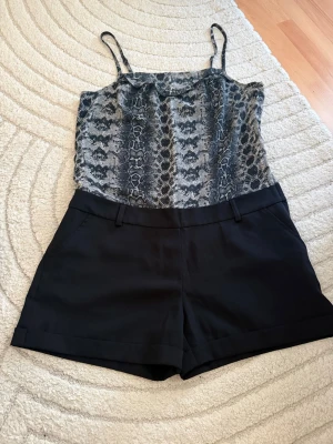 Jumpsuit med ormmönstrad topp - Snygg jumpsuit med smala axelband, ormmönstrad topp i grått och svart samt svarta shorts nertill. Perfekt för dig som gillar trendiga mönster och vill ha en cool look. Toppdelen är lätt och luftig, shortsdelen har fickor och bälteshällor.
