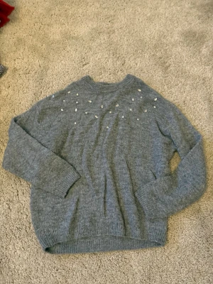 Grå stickad tröja med pärlor H&M - Mjuk grå stickad tröja från H&M med pärldetaljer över bröstet och axlarna. Tröjan har rund halsringning, långa ärmar och ribbade muddar vid ärmslut och nederkant. Barnstorlek men ca storlek XS eller S i vanlig.