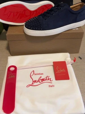 Blåa sneakers från Christian Louboutin - Snygga blåa sneakers från Christian Louboutin med klassisk röd sula och coola nitar på tån. Skorna är låga och gjorda i mocka med vita gummisulor. Perfekt för dig som vill sticka ut med exklusiva detaljer och ikonisk design.