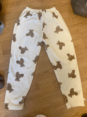Vita mjukisbyxor med nallemönster - Supergosiga vita loungewear-byxor med bruna nallar över hela tyget. Byxorna har resår i midjan och är gjorda i ett fluffigt, fleece-liknande material som är perfekt för mysiga hemmadagar.
