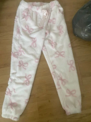 Vita fluffiga loungewear-byxor med rosetter - Supermjuka och mysiga vita loungewear-byxor med ljusrosa rosett-mönster och satinband i midjan. Perfekta för chill hemma eller sleepovers. Byxorna har resår i midjan och är gjorda i ett fluffigt material som känns riktigt skönt mot huden.