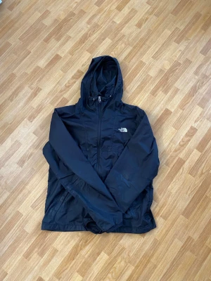 Svart vindjacka från The North Face - Snygg svart vindjacka från The North Face med huva och dragkedja framtill. Jackan har två sidofickor med dragkedja och jackan är enkel att packa ner i väskan. Den passar bra till någon som är S-M