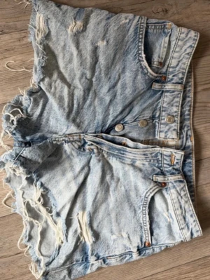 Ljusa jeansshorts med fransar - Säljer ett par snygga ljusblå jeansshorts med  fransiga kanter och slitna detaljer. Shortsen har hög midja, klassiska fem fickor och knappar framtill. Perfekta för sommaren och ger en avslappnad stil dessutom är dom väldigt bekväma!!💕💕