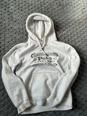 Grå hoodie från Polo Ralph Lauren - Snygg ljusgrå hoodie från Polo Ralph Lauren med svart tryck framtill. Klassisk modell med huva och dragsko, samt en stor magficka. Tillverkad i mjukt bomullsmaterial som är skönt mot huden. Perfekt för en avslappnad och trendig look.