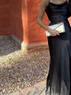 Svart bal långklänning med smala axelband - Elegant svart långklänning med smala axelband och öppen rygg. Klänningen har en figurnära passform och är tillverkad i ett glansigt material som ger en lyxig känsla. Perfekt för bal eller festliga tillfällen. Storlek S 
