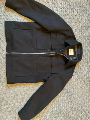 Svart overshirt från Jack and Jones  - Stilren svart overshirt från Jack and Jones med två stora bröstfickor och dragkedja framtill. Overshirten har klassisk krage och är tillverkad i ett tjockare material som ger en schysst struktur. Perfekt lager-på-lager-plagg för en clean look.