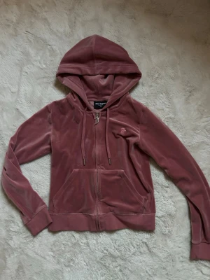 Rosa velourhoodie från Juicy Couture - Mysig rosa hoodie i velour från Juicy Couture med dragkedja, huva och kängurufickor. Klassisk logga broderad på bröstet och justerbara snören i huvan. Perfekt för en avslappnad och trendig look.