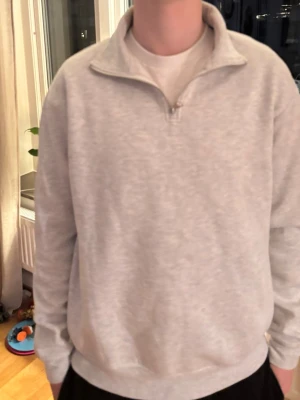 Ljusgrå halvzip sweatshirt - Ljusgrå sweatshirt med halv dragkedja och hög krage. Tröjan har en avslappnad passform och är tillverkad i mjukt material, perfekt för en chill och stilren look. Långärmad modell som passar året runt. Har några små fläckar som syns på bilderna