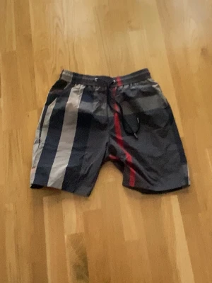 Burberry shorts - Säljer ett par snygga badbyxor med breda vertikala ränder i svart, grått, vitt och rött. De har elastisk midja med dragsko och en bakficka. Perfekta för stranden eller poolen.