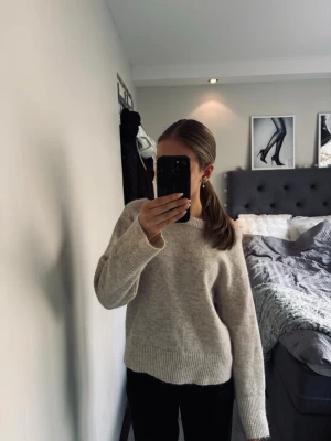 Beige stickad tröja  - Mysig beige stickad tröja och urringning vid halsen. Breda ribbade muddar vid ärmslut och nederkant. Tröjan har en avslappnad passform och är tillverkad i en mjuk ullblandning, perfekt för kyliga dagar.