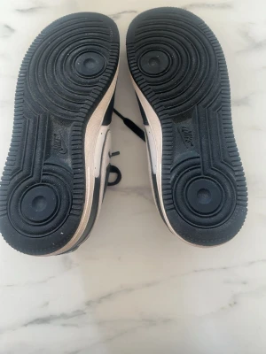 Nike Air Force 1 svartvita sneakers - Svartvita Nike Air Force 1 sneakers med klassisk låg siluett. Skorna har vit tåbox med perforeringar, svarta paneler och vit Swoosh på sidan. Yttersulan är svart med tydlig Air-dämpning. Perfekta för en streetwear-look.