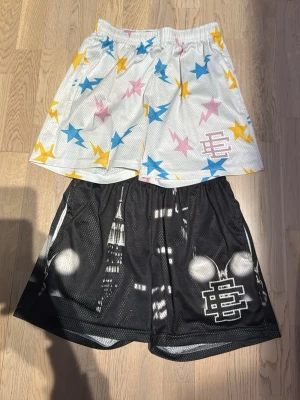 EE shorts - Säljer två par basketinspirerade shorts i mesh. Ena paret är vita med stjärnmönster i blått, rosa och gult, det andra är svarta med stadsmotiv och vit logga. Båda har resår i midjan som och ledig passform. Perfekta för en sportig look. 150kr styck om man köper båda för man dem för 250kr!!!