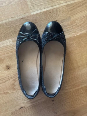 Svarta ballerinaskor med lackdetalj - Snygga svarta ballerinaskor med glansig lackad tå och dekorativ rosett framtill. Ovandelen har en textilstruktur som ger en cool kontrast till lacken. Perfekta för dig som gillar klassisk men trendig stil. Har sko skavs skydd i. Storlek 37 men passar mer 38