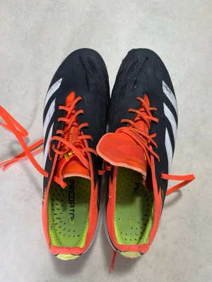 Adidas Predator fotbollsskor svart/orange - Snygga Adidas Predator fotbollsskor i svart med vita detaljer och orange snörning. Skorna har låg profil, mönstrad ovandel och fasta dobbar för spel på gräs. Insidan är limegrön och märket syns tydligt på sidan. Perfekta för dig som vill sticka ut på planen.