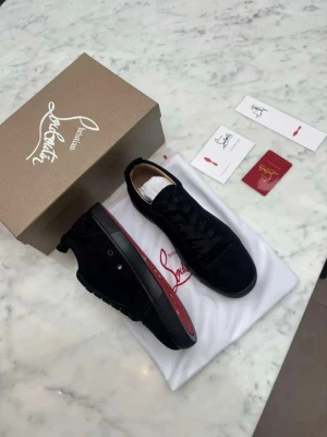(Äkta) Christian Louboutin sneakers - Snygga svarta sneakers från Christian Louboutin med klassisk röd sula och svarta skosnören. Skorna är låga och har en stilren design i mocka och läder. Perfekta för dig som vill sticka ut med exklusiva detaljer och ikonisk look.