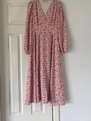 Blommig långklänning från Neo Noir - Långklänning från Neo Noir med blommigt mönster i rött, rosa och vitt. Klänningen har v-ringning, långa ärmar och en luftig passform. Perfekt för vår och sommar med sitt lätta material och romantiska stil.
