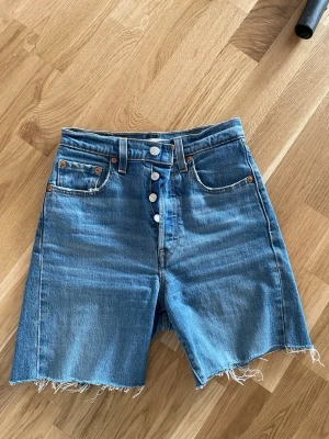 Levi's Ribcage denimshorts blå - Snygga blå denimshorts från Levi's Ribcage med hög midja och raka ben. Shorts med råa, fransiga kanter och klassiska fem fickor. Knappar framtill och cool vintagekänsla. Perfekta för sommaren!