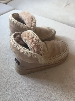 Beige fårskinnstofflor från Mou, stl 38,5 - Mysiga beige tofflor från Mou med mjukt fårskinnsfoder och dekorativa stickade detaljer längs kanterna. Tofflorna har en högre modell och är perfekta för att hålla fötterna varma. Utsidan är i mocka och insidan är fluffig och skön.
