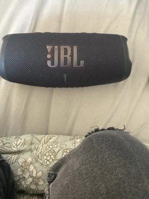 JBL svart trådlös högtalare - Snygg svart trådlös högtalare från JBL med robust tygmaterial och tydlig JBL-logga i metall på framsidan. Cylindrisk form med gummiklädda ändar och smidiga knappar för Bluetooth, volym och power på ovansidan. Perfekt för att ta med överallt och spela musik trådlöst.