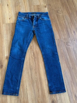  Nudie Jeans grim Tim  - Säljer ett par klassiska blå jeans från Nudie Jeans Co i storlek 31/30. Jeansen har raka ben, fem fickor och snygga kontrastsömmar. Materialet är slitstark denim i bomull och modellen har knappgylf. Perfekt för dig som gillar stilrena och tidlösa jeans.