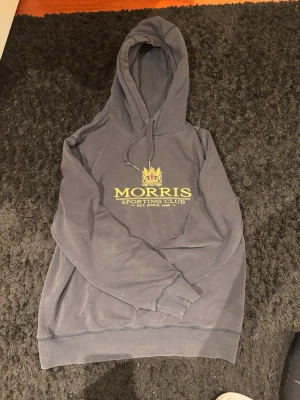 Svart hoodie från Morris Sporting Club - Navy hoodie från Morris Sporting Club med broderad logga och emblem i guld på bröstet. Klassisk modell med huva och dragsnören, ribbade muddar och mjukt material. Perfekt för en avslappnad och stilren look.