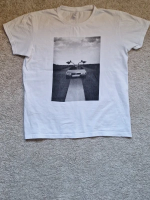 Vit t-shirt med biltryck - Vit t-shirt från Fruit of the Loom med ett svartvitt tryck av en bil med öppna dörrar på framsidan. Klassisk rund hals och korta ärmar. Perfekt för dig som gillar coola och unika motiv. Storlek XS