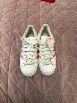 Adidas Superstar vita med rosa ränder - Säljer ett par klassiska Adidas Superstar sneakers i vitt läder med rosa metallic-ränder och matchande hälflik. Skorna har den ikoniska snäckformade tån och vita skosnören. Perfekta för dig som vill ha en clean och sportig look med en touch av färg.