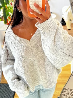 Ljusgrå kabelstickad tröja - Säljer en ljusgrå kabelstickad tröja med bred v-ringning och oversized passform. Tröjan har långa ärmar och ribbade muddar vid ärmslut och nederkant. Perfekt för att skapa en chill och trendig look. Materialet är mjukt och mysigt, troligtvis akryl eller ullblandning.