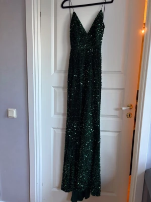 Grön glittrig långklänning med smala band - Snygg grön långklänning med smala axelband och djup v-ringning. Klänningen är täckt av glittriga paljetter och har en elegant passform som framhäver siluetten. Perfekt för dig som vill sticka ut på bal eller fest.