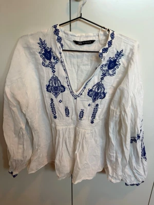 Vit blus med blå broderier från Zara - Snygg vit blus från Zara med blå broderade detaljer och v-ringning. Blusen har långärmade puffiga ärmar och är tillverkad i ett lätt och luftigt material, perfekt för en cool och avslappnad look. Broderierna ger en unik och bohemisk känsla.